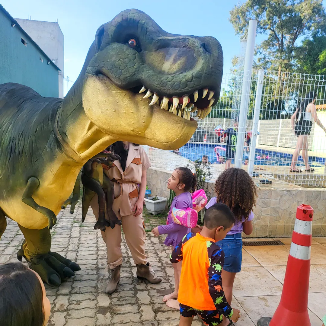 Ator caracterizado de explorador ao lado de réplica hiper-realista de dinossauro T-Rex gigante em festa infantil.